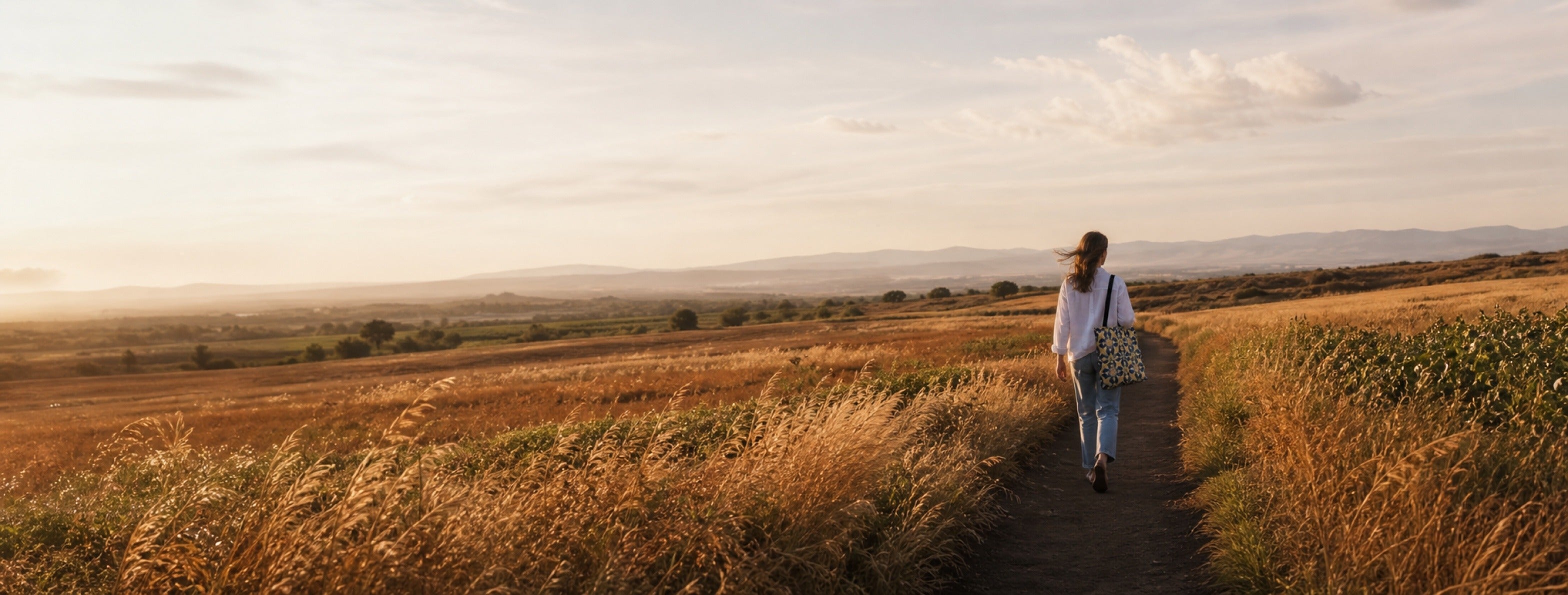 Woman walking a red-earth field path carrying a Zellige Woven Tote