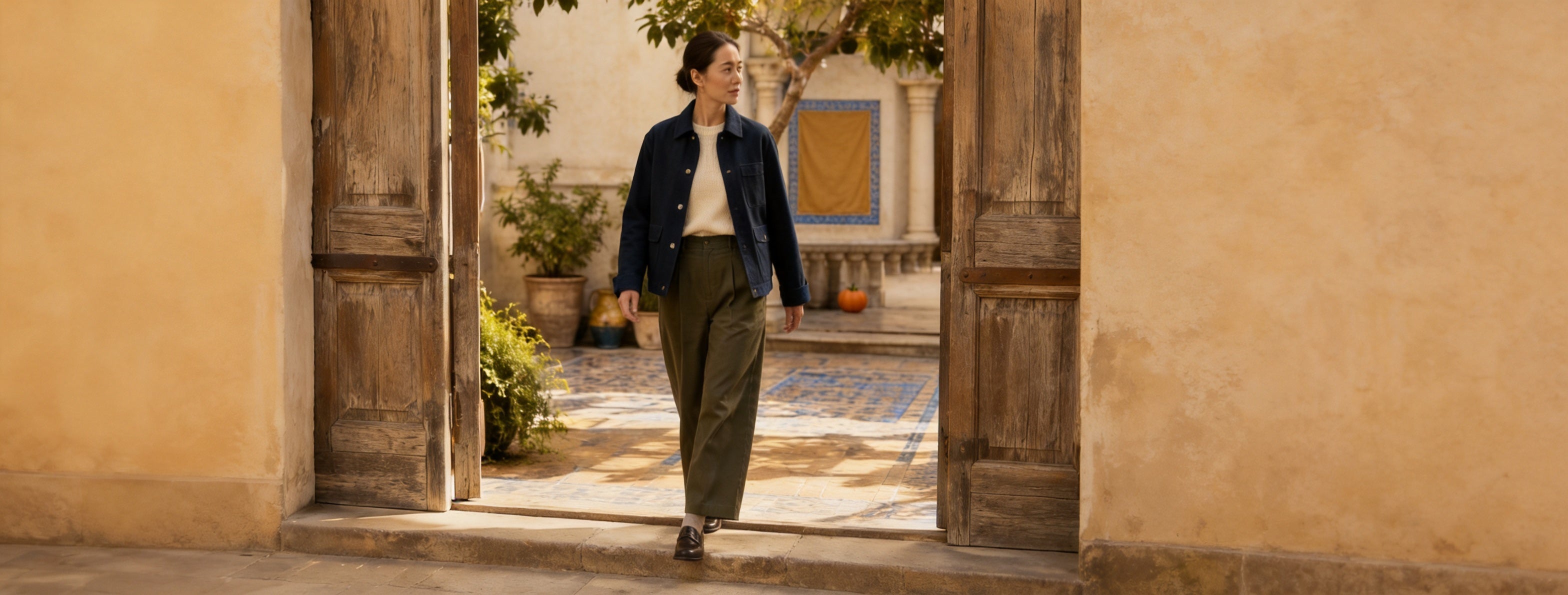 A woman walking through a sunlit stone threshold toward a quiet garden path.